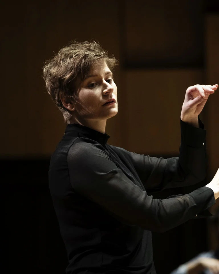 Anja Bihlmaier: A conductor leads an orchestra against a blurred background. She raises her baton in a dynamic gesture.
