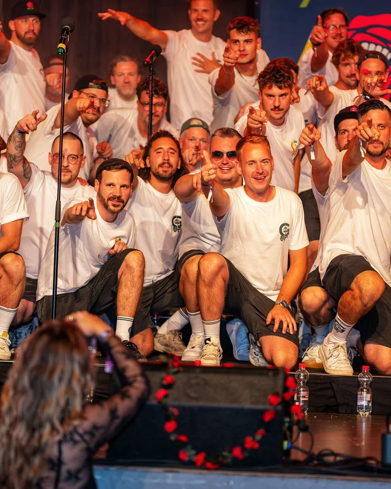 Eine Gruppe von Menschen steht auf einer Bühne und trägt weiße Hemden. Sie lachen und zeigen in die Kamera.