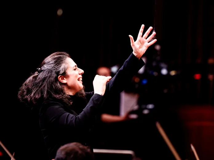 Eine Dirigentin leitet mit energischem Ausdruck ein Orchester. Musiker konzentrieren sich auf ihre Instrumente im Hintergrund.