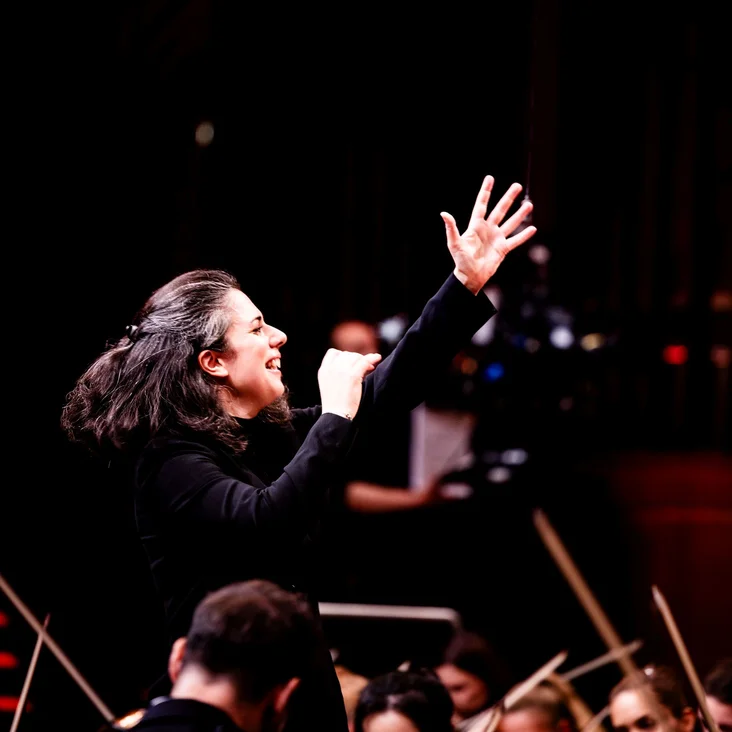 Eine Dirigentin leitet mit energischem Ausdruck ein Orchester. Musiker konzentrieren sich auf ihre Instrumente im Hintergrund.