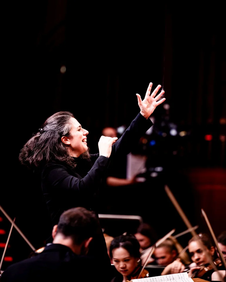 Eine Dirigentin leitet mit energischem Ausdruck ein Orchester. Musiker konzentrieren sich auf ihre Instrumente im Hintergrund.