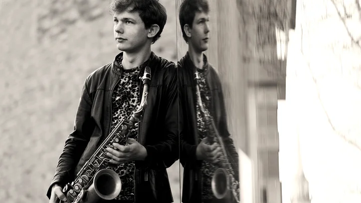 Ein junger Mann lehnt an einer Wand und hält ein Saxophon, dessen Spiegelbild im Fenster zu sehen ist. Sepia-Ton.