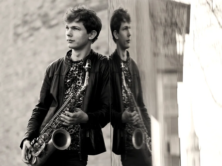 Ein junger Mann lehnt an einer Wand und hält ein Saxophon, dessen Spiegelbild im Fenster zu sehen ist. Sepia-Ton.