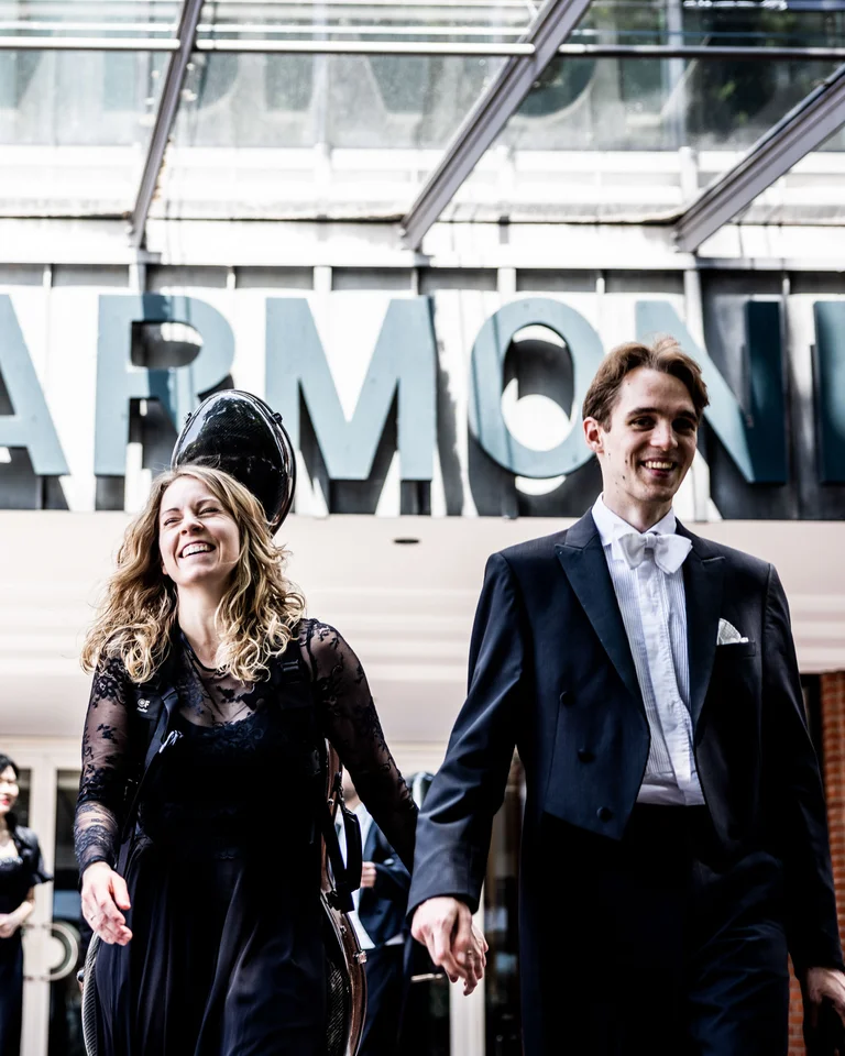 Mann und Frau in formeller Kleidung vor einem Gebäude mit dem Schriftzug 'PHILHARMONIE'. Sie lächeln und gehen nach draußen.
