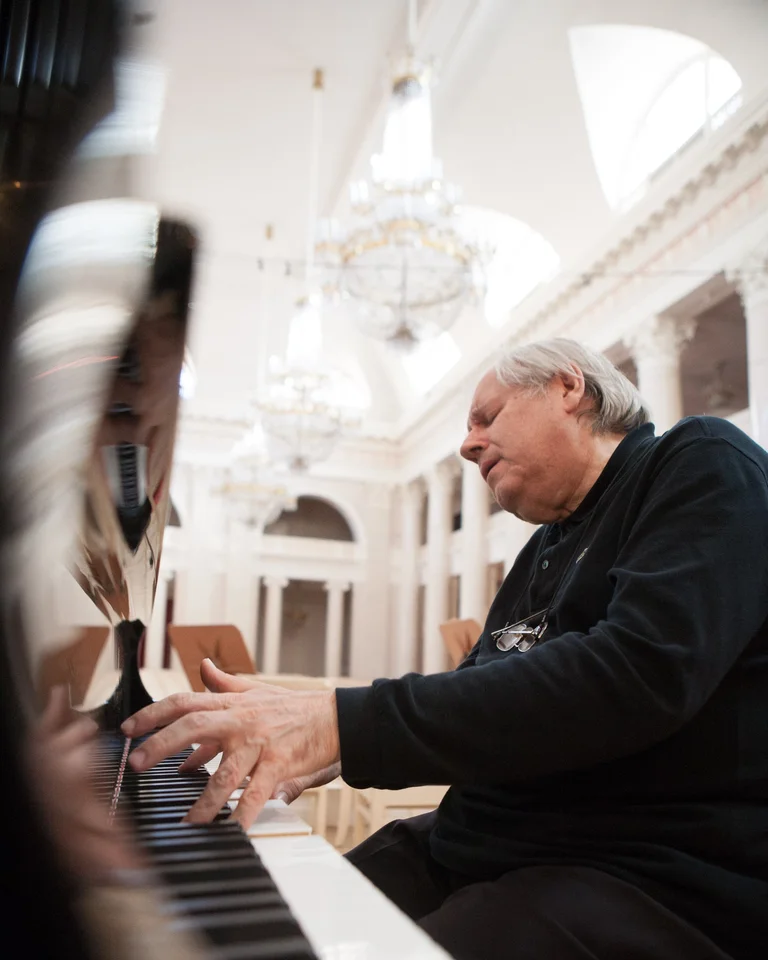 Ein älterer Mann (Grigory Sokolov) spielt Klavier in einem eleganten, hell erleuchteten Saal mit großen Kronleuchtern und hohen Bögen.