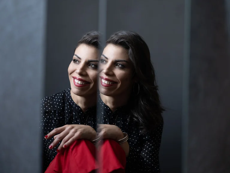 Eine lächelnde Frau sitzt nahe an einem Spiegel und trägt rote und schwarze Kleidung. Ihr Spiegelbild ist zu sehen.