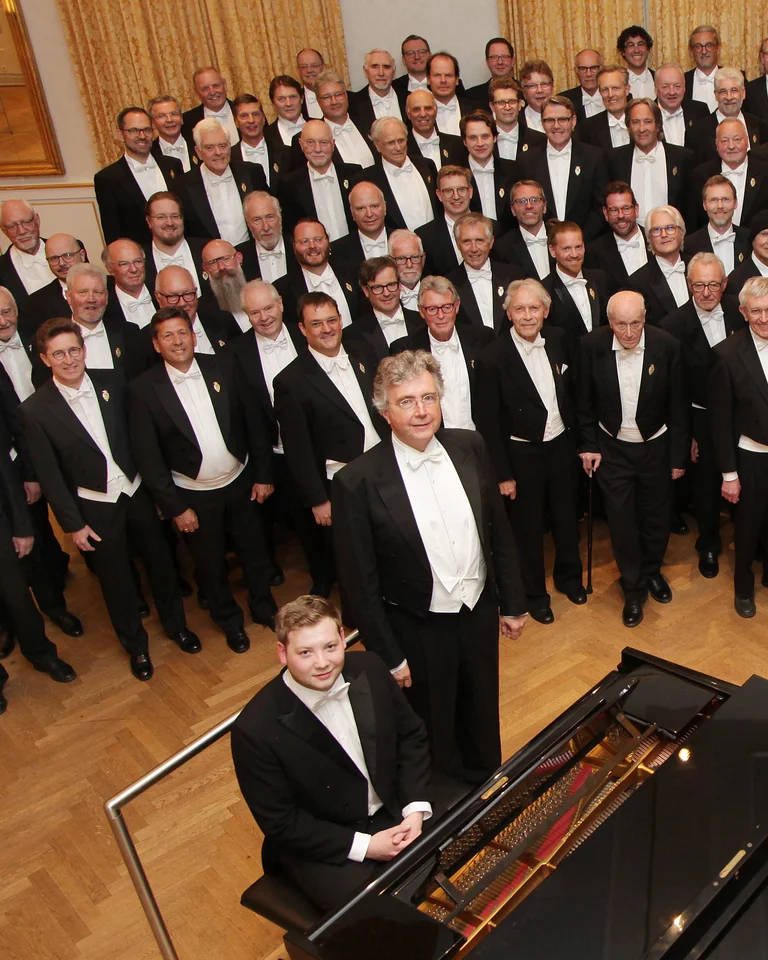 Ein großer Männerchor in Frack steht in einem Konzertsaal vor einem Flügel. Zwei Dirigenten stehen vorne.
