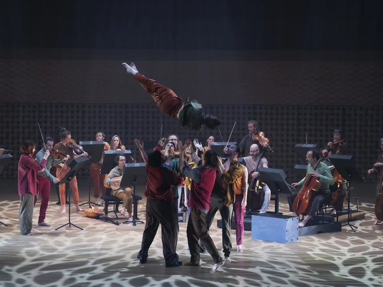 Ein Ensemble von Musikern spielt live, während Akrobaten in der Luft kunstvolle Bewegungen ausführen. Die Bühne ist stimmungsvoll beleuchtet.