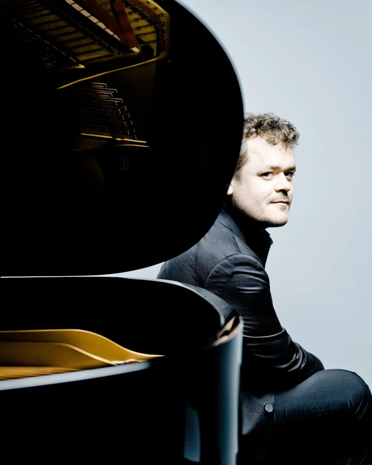 Ein Mann sitzt neben einem Flügel, stilvoll in Schwarz gekleidet. Die Atmosphäre ist modern und elegant.