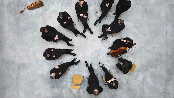 Zwölf Musiker in schwarzer Kleidung (Die 12 Cellisten der Berliner Philharmoniker) sitzen im Kreis auf grauem Boden; Streichinstrumente wie Celli sind vorhanden.