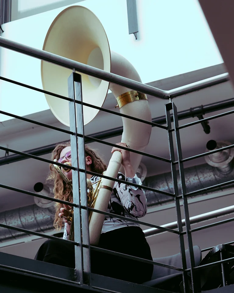 Eine Person spielt ein großes Blechblasinstrument, möglicherweise eine Tuba, auf einem Balkon in einem modernen Gebäude.