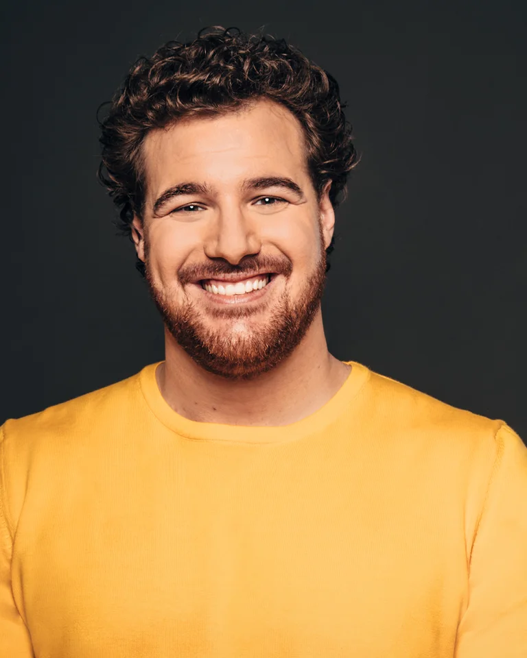 Ein Mann mit lockigem Haar und Bart lächelt freundlich. Er trägt ein gelbes Shirt. Der Hintergrund ist dunkel.