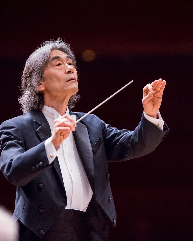 Ein Dirigent (Kent Nagano) im schwarzen Anzug leitet ein Orchester mit lebhaften Bewegungen seines Taktstocks auf einer Konzertbühne.