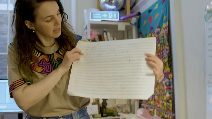 Anna Meredith is holding a sheet of music paper with handwritten musical notation. Colorful fabrics and shelves are visible in the background.