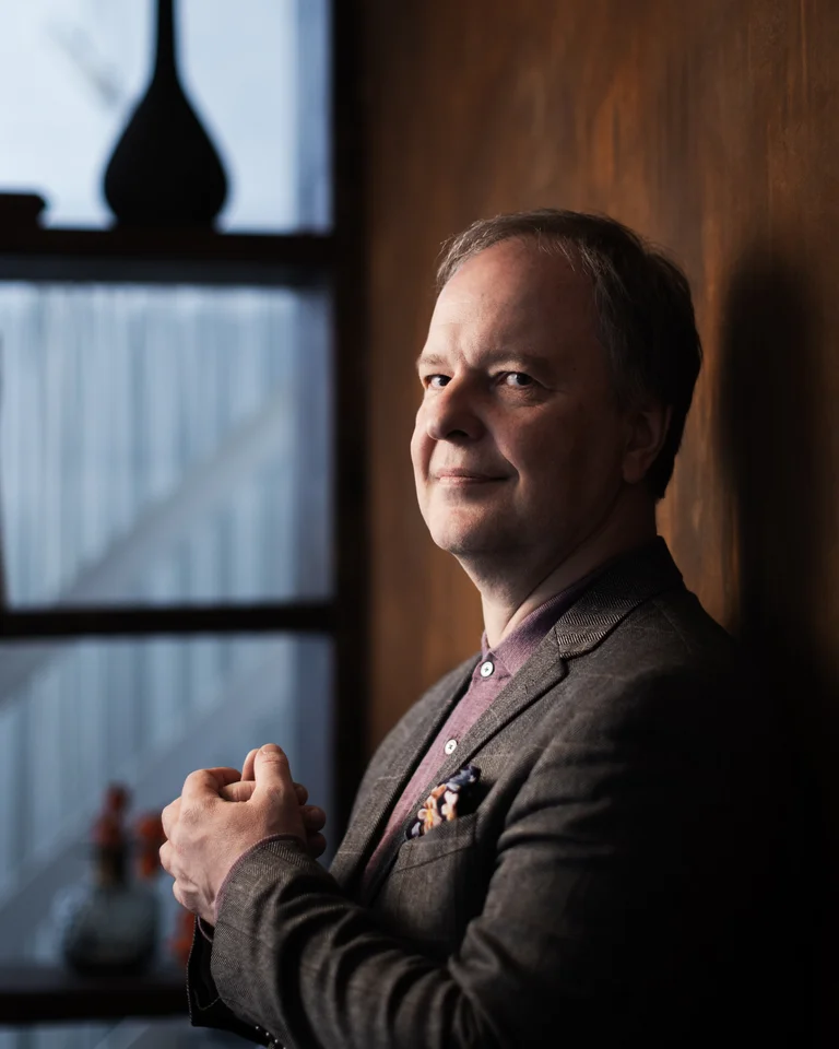 Ein Mann in einem Anzug steht vor einem Bücherregal. Im Hintergrund sind eine alte Nähmaschine und dekorative Gegenstände.