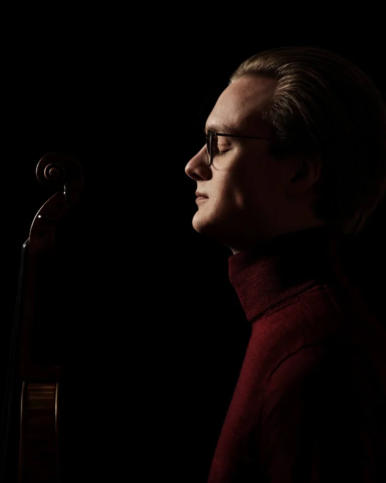 Ein Mann in einem dunklen Raum, von der Seite beleuchtet, steht neben einem Streichinstrument. Er trägt einen Rollkragenpullover.