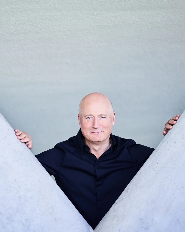 Paavo Järvi: Ein Mann in schwarzer Kleidung steht zwischen zwei großen, grauen Wänden, die ein V formen. Er blickt direkt in die Kamera.