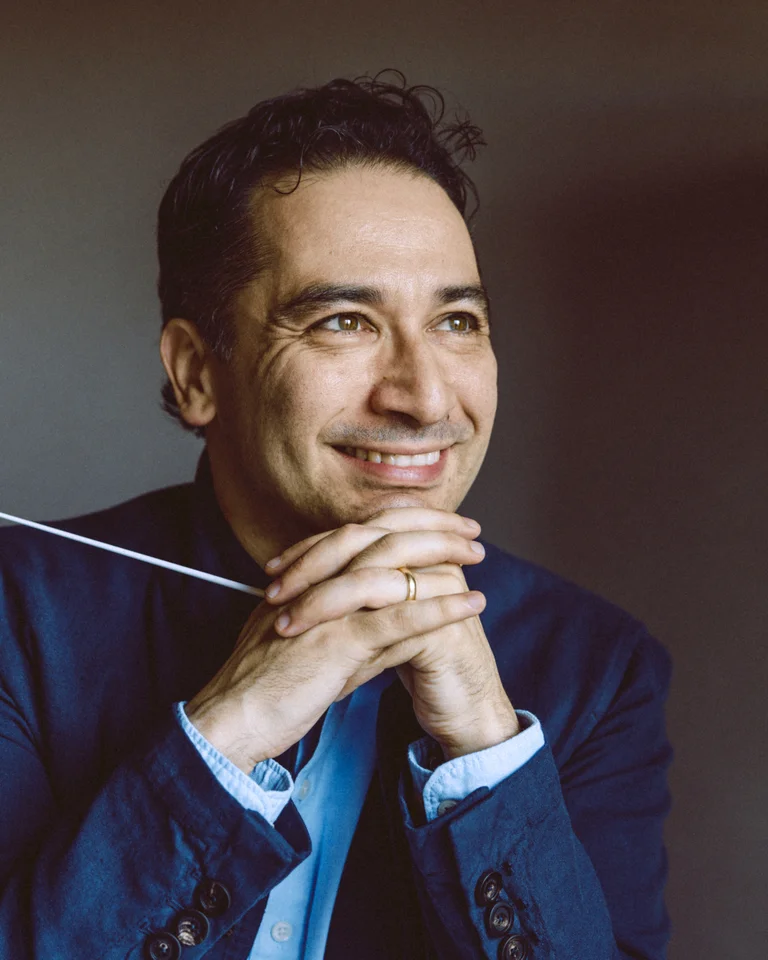 Ein lächelnder Mann im Anzug hält einen Dirigentenstab in den Händen, als ob er ein Orchester leitet.