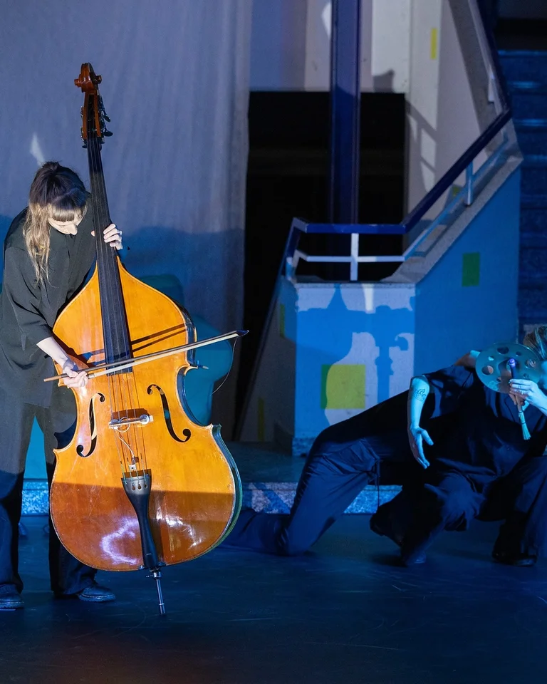 Eine Musikerin spielt Kontrabass auf einer Bühne. Neben ihr tanzt eine Person, die in blaues Licht getaucht ist.