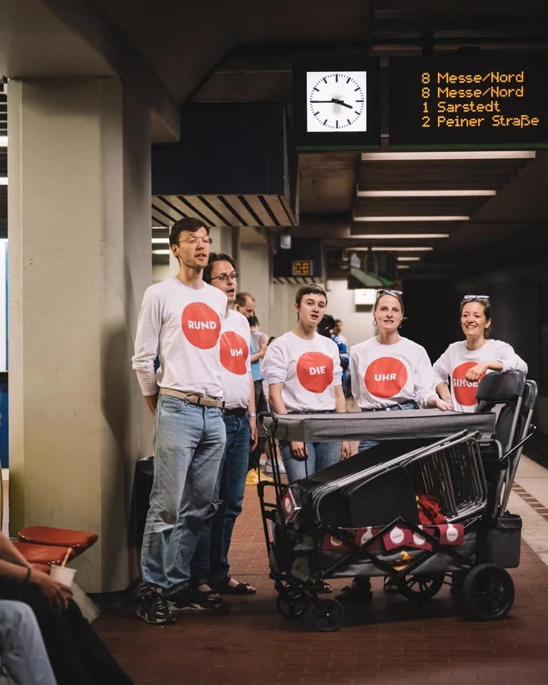 A choir (ENSEMBLE SOZUSINGEN) sings on a subway platform, surrounded by waiting passengers. The members wear white T-shirts with red prints.