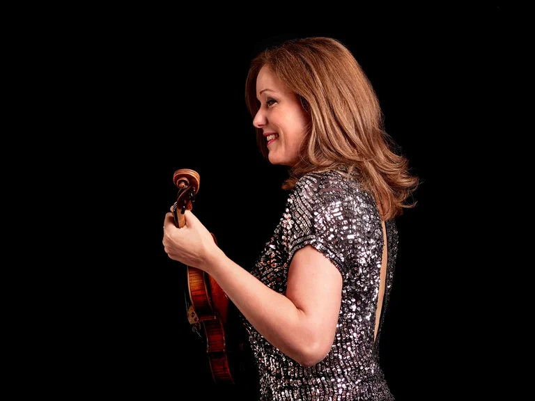 Frau (Isabelle van Keulen) lächelt und hält eine Geige in der Hand. Sie trägt ein gemustertes Kleid vor einem schwarzen Hintergrund.