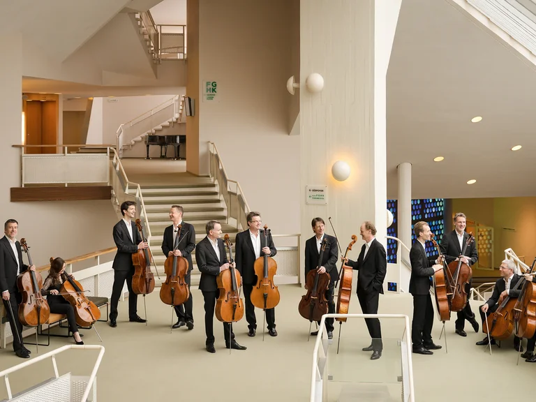 Eine Gruppe von Musikern mit Celli steht in einem modernen Gebäude (Die 12 Cellisten der Berliner Philharmoniker), bereit für eine Aufführung. Lichte, offene Architektur im Hintergrund.