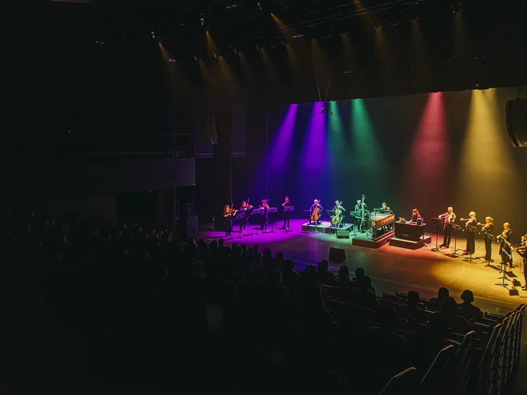 Eine Bühne in einem dunklen Saal mit musikalischen Darstellern, beleuchtet von bunten Lichtern in Blau, Lila, Grün und Gelb.