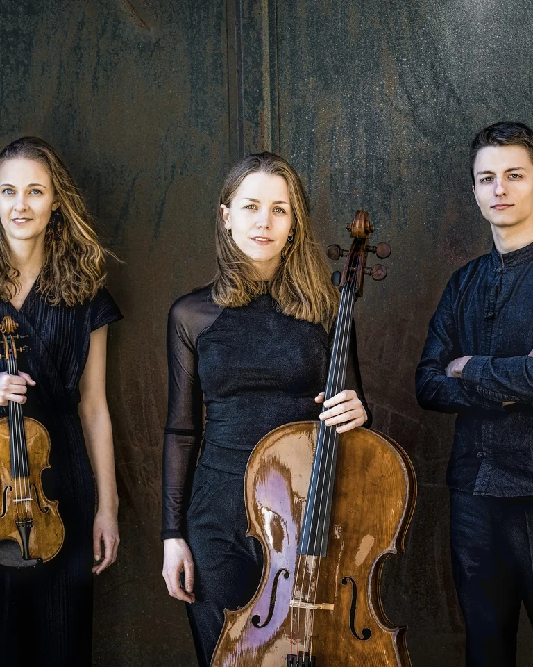 Die drei Mitglieder des Amelio Trio posieren, teils mit ihren Instrumenten, vor einem dunklen Hintergrund. Sie tragen schwarze Konzertkleidung und blicken in die Kamera.