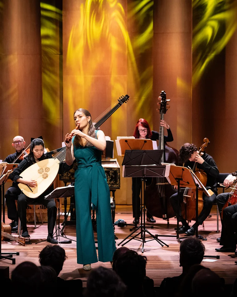 Ein Orchester spielt auf einer Bühne mit goldenen Lichtern im Hintergrund. Eine Flötistin steht im Vordergrund (Lucie Horsch mit dem Orchestra of the Eighteenth Century).