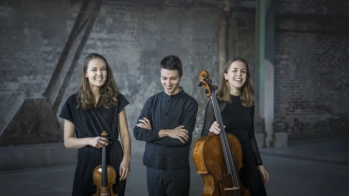 Drei Musiker (Amelio Trio) stehen in einem industriellen Raum. Zwei halten Streichinstrumente, ein Geige und ein Cello. Sie tragen dunkle Kleidung.
