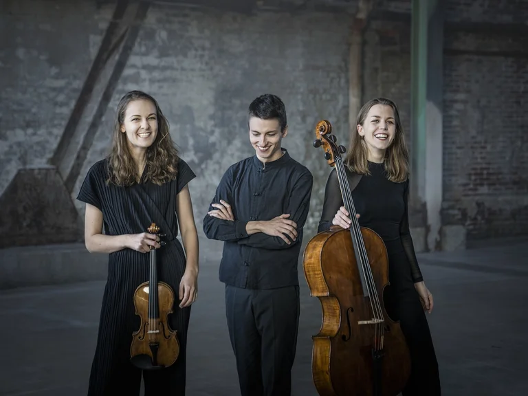 Drei Musiker (Amelio Trio) stehen in einem industriellen Raum. Zwei halten Streichinstrumente, ein Geige und ein Cello. Sie tragen dunkle Kleidung.