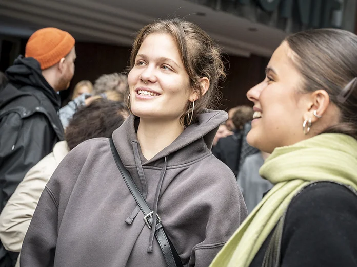 Two women smiling outside the entrance of the Philharmonie. Several people gather around them, waiting to go in.