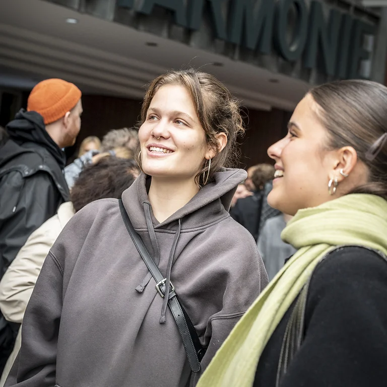 Two women smiling outside the entrance of the Philharmonie. Several people gather around them, waiting to go in.