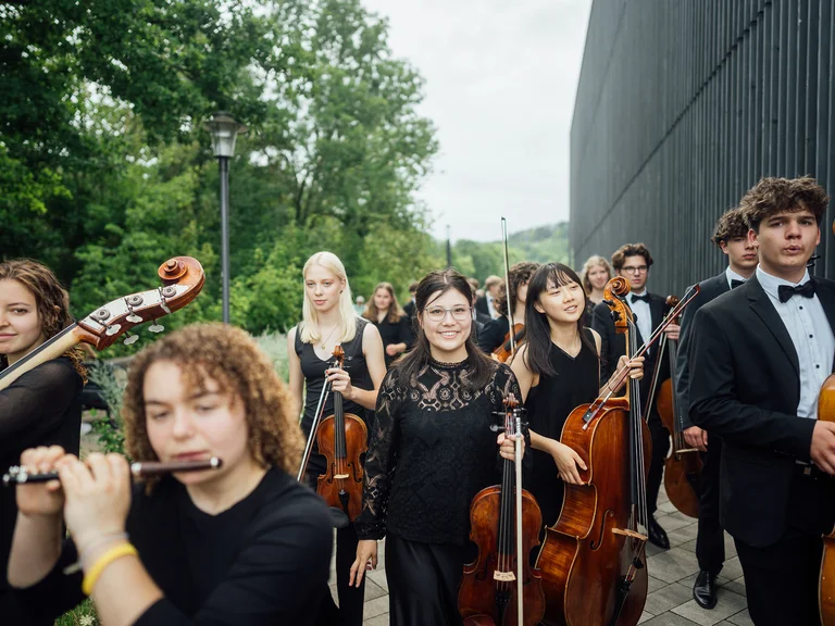 Eine Gruppe junger Musiker (Bundesjugendorchester) in formeller Kleidung mit verschiedenen Instrumenten, wie Violoncelli und einer Flöte, draußen bei einem Konzert.