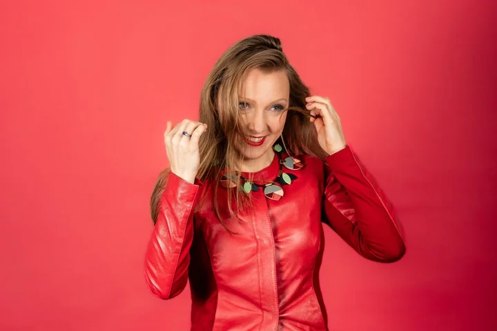 Eine Frau (Olga Pashchenko) in einer roten Lederjacke steht vor einem roten Hintergrund. Sie trägt eine bunte Halskette und lächelt.