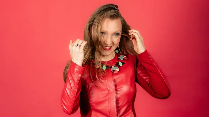 Eine Frau (Olga Pashchenko) in einer roten Lederjacke steht vor einem roten Hintergrund. Sie trägt eine bunte Halskette und lächelt.