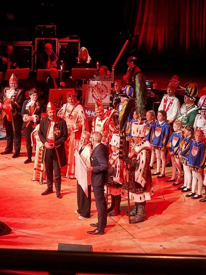 Menschen in traditionellen Kostümen und Uniformen auf einer Bühne, beleuchtet von roten Lichtern, bei einem Karnevalsereignis.