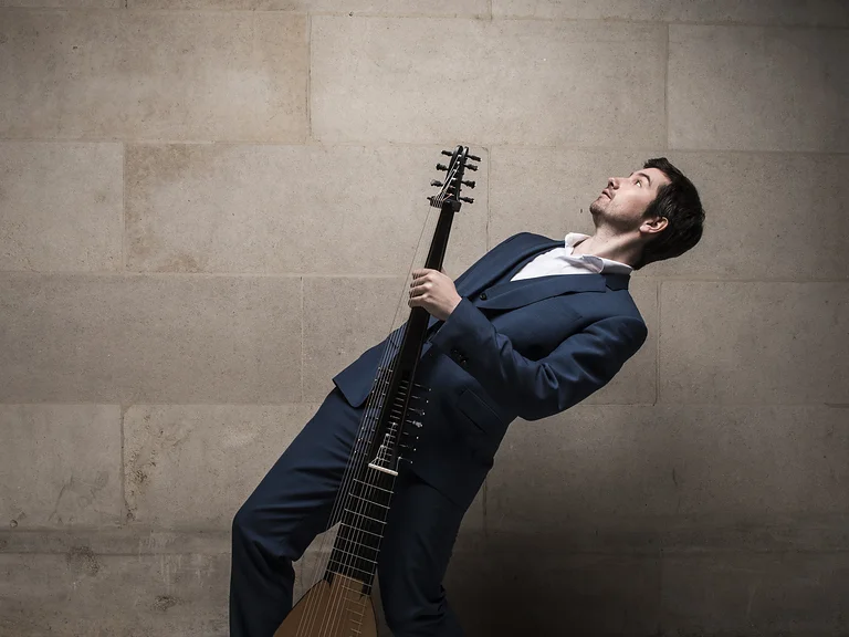 Ein Mann (Thomas Dunford) im Anzug lehnt sich zurück und hält einer Laute, vor einer Steinwand.