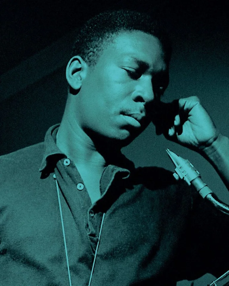 Ein Mann steht in einem Raum, spricht in ein Telefon. Neben ihm steht ein Saxophon. Das Bild hat einen blauen Farbton.