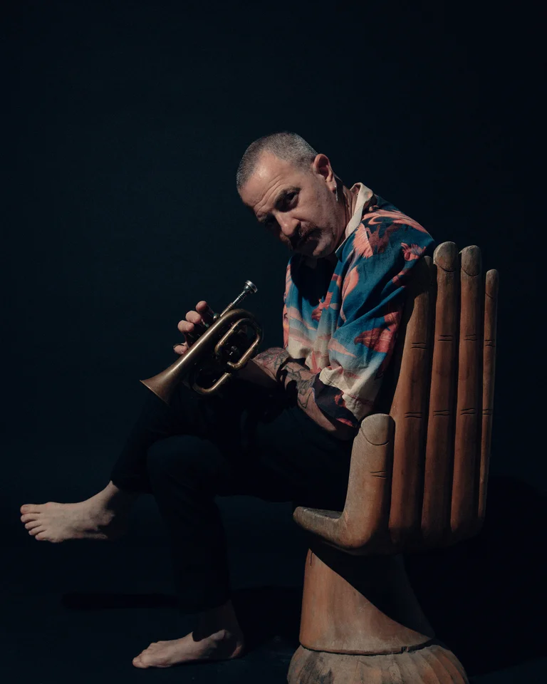 Ein Mann mit einer bunten Jacke (Avishai Cohen) sitzt barfuß auf einem hölzernen Handstuhl und spielt Trompete in einem dunklen Raum.