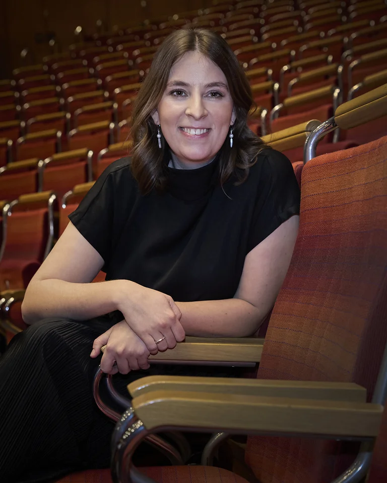 Charlotte Oelschlegel Frau sitzt lächelnd in einem leeren Konzertsaal. Sie trägt ein schwarzes Outfit und sitzt auf einem der roten Sitze der Kölner Philharmonie.