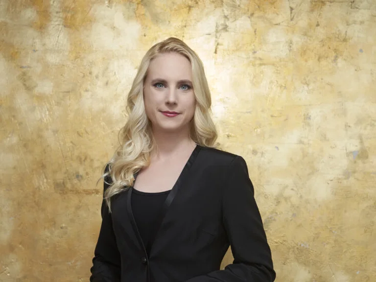 Eine Frau in einem schwarzen Anzug hält einen Taktstock vor einem goldenen Hintergrund.