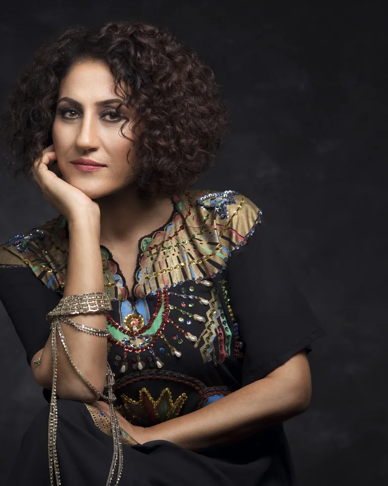 Eine Frau mit lockigem Haar, die einen verzierten, ethnisch inspirierten Umhang trägt, lehnt ihren Kopf nachdenklich auf ihre Hand.