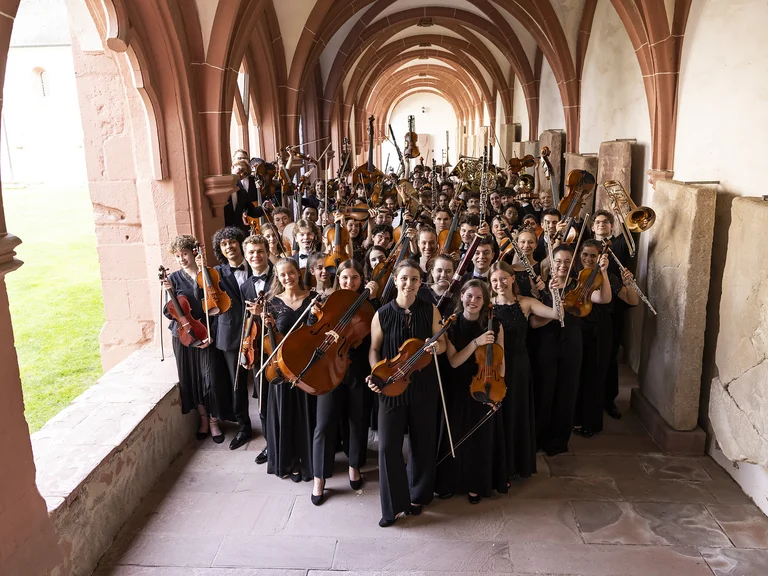 Eine große Gruppe von Musikern (Bundesjugendorchester) in schwarzen Outfits steht in einem historischen, gewölbten Gang mit Bögen, jede Person hält ein Instrument.