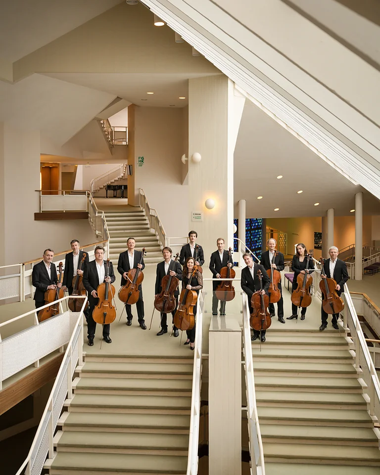 Eine Gruppe von Musikern (Die 12 Cellisten der Berliner Philharmoniker) mit Streichinstrumenten steht auf einer modernen Treppe in einem eleganten Veranstaltungsgebäude.