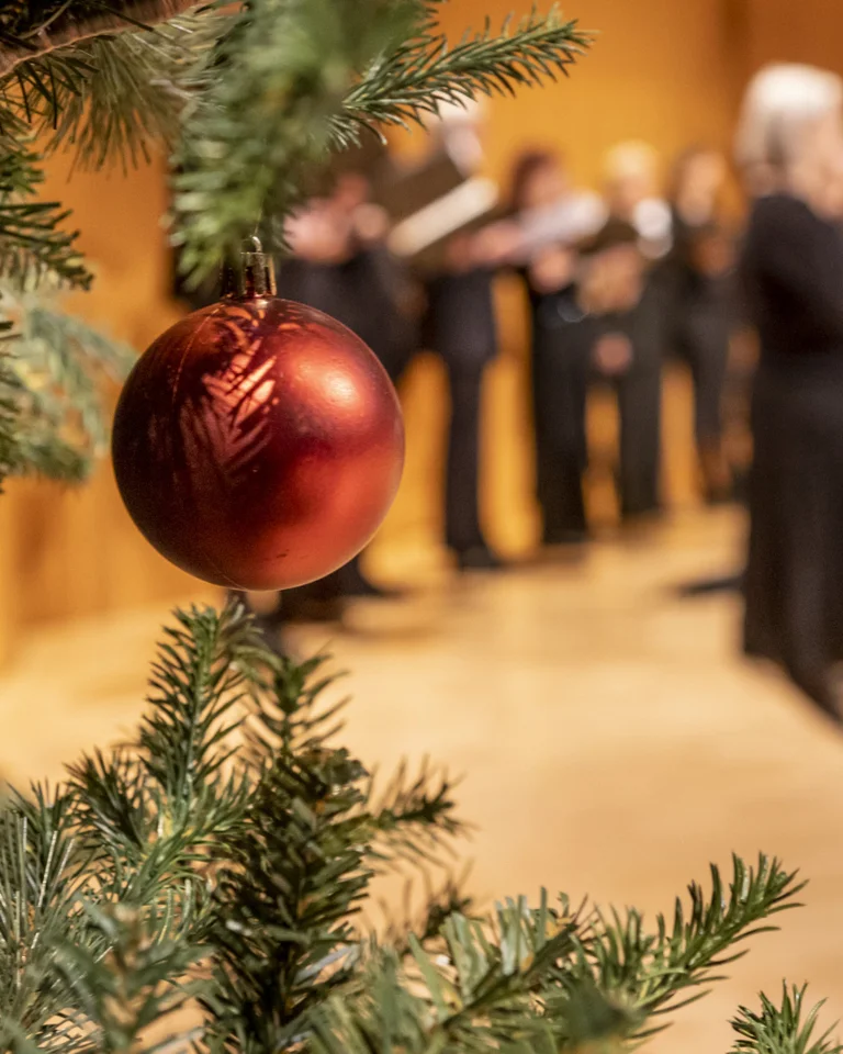 Nahaufnahme einer roten Weihnachtskugel an einem Baum. Im Hintergrund singt ein Chor in festlicher Atmosphäre.