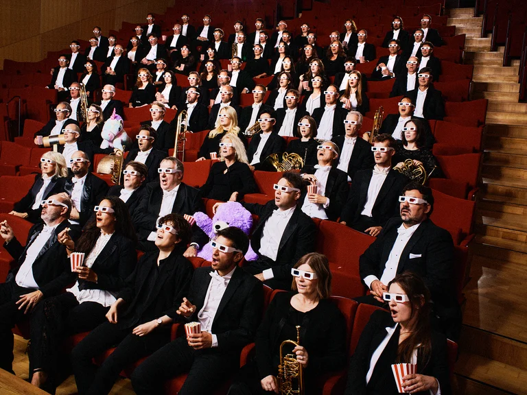Ein Orchester in Konzertkleidung sitzt in einem Auditorium. Einige halten Blasinstrumente und tragen Blumenhüte oder Masken.