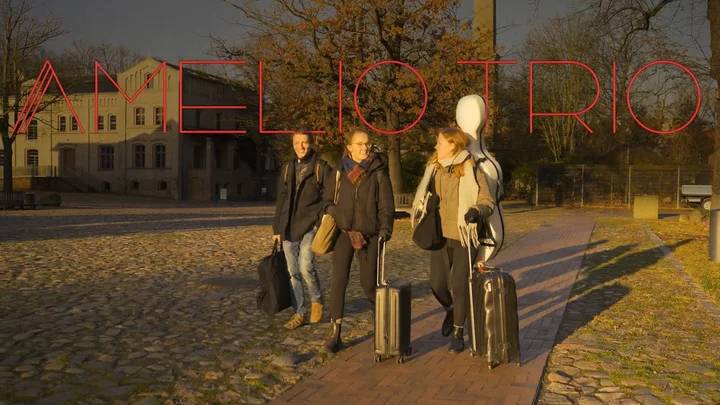 Die Mitglieder des Amelio Trios gehen mit Trolleys und Instrumentenkoffern eine gepflasterte Straße bei Sonnenuntergang entlang.