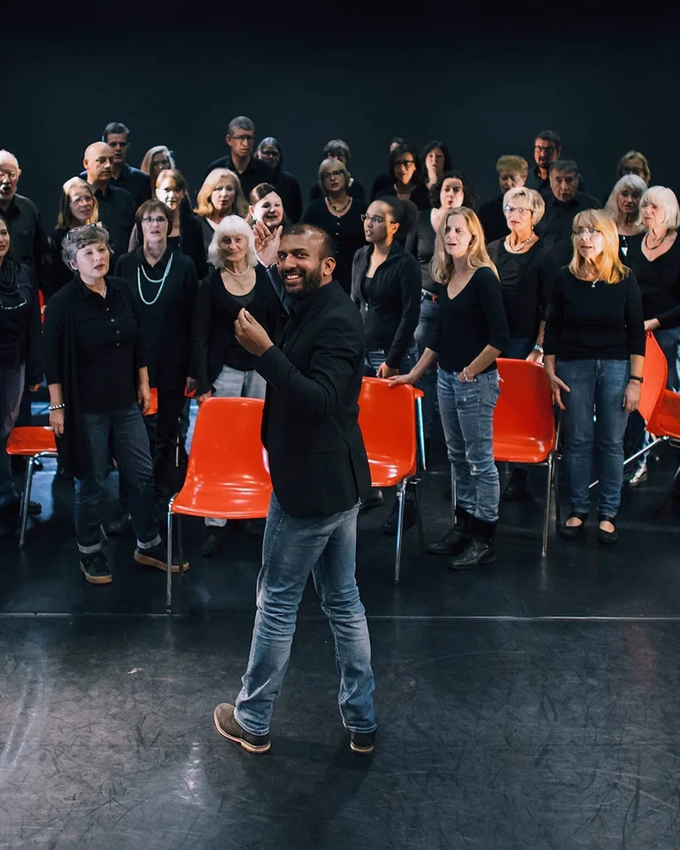 A large group of singers on stage. They wear black tops and jeans, some stand while others sit on red chairs.