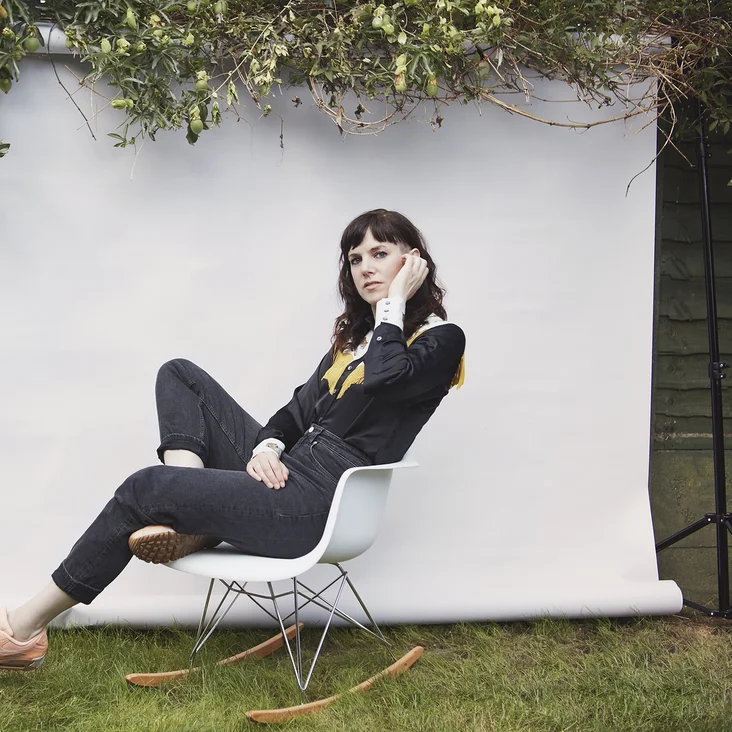 Eine Frau (Anna Meredith) sitzt lässig auf einem Stuhl vor einem neutralen Hintergrund im Freien, umgeben von Gräsern und grünen Zweigen.